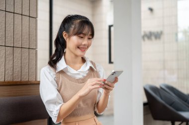 Genç ve güzel bir Asyalı kadın binada oturuyor ve akıllı telefonunu kullanıyor. Sohbet etmek, telefonda kaymak, mobil uygulama kullanmak, online alışveriş, mesajları okumak