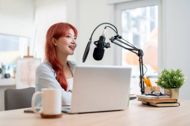 Kızıl kıvırcık saçlı güzel bir Asyalı kadın podcaster stüdyosunda çalışıyor, mikrofona konuşuyor ve şovu için ilgi çekici hikayeler anlatıyor..