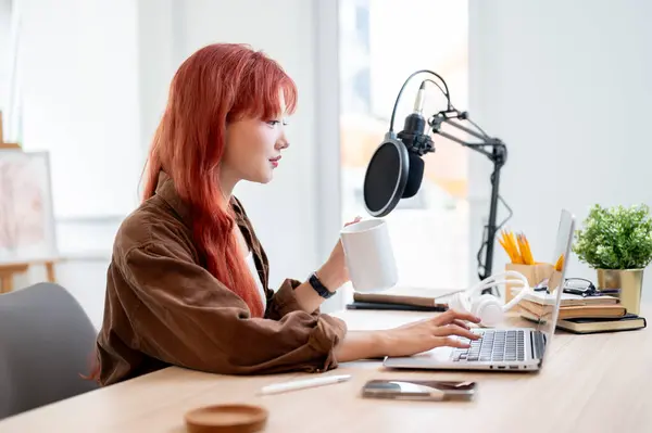 stock image A confident and beautiful Asian female podcaster is working in her home studio, enjoying a coffee while recording a new episode of her podcast.