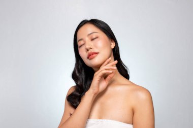 Göz kamaştırıcı bir yüze ve bronzlaşmış tene sahip genç bir Asyalı güzel gri bir arkaplana karşı poz veriyor, yüzüne nazikçe dokunuyor gözleri kapalı, rüya gibi bir ifadede kayboluyor. yüz tedavisi kozmetik