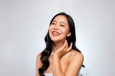 Parlak tenli ve uzun siyah saçlı genç bir Asyalı güzellik modeli gri bir arka planda poz veriyor, gülümsüyor, dişlerini gösteriyor ve nazikçe yüzüne dokunuyor. yüz tedavisi, makyaj, kozmetik