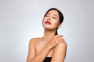 Parlayan, sağlıklı bronzlaşmış tenli, muhteşem bir Asyalı güzellik modeli gri bir stüdyo arka planında gözleri kapalı tenine dokunarak poz veriyor. tedavi, spa, makyaj, kozmetik, plastik cerrahi