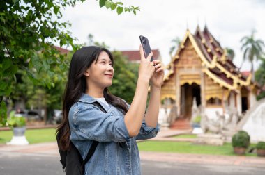 Kot ceketli mutlu, güzel bir Asyalı kadın gezgin akıllı telefonuyla Tayland tapınağının fotoğrafını çekiyor, yalnız gezisi sırasında bir tapınağı ziyaret ediyor. Yaşam tarzları, kablosuz teknoloji, seyahat