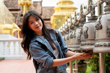 Tayland 'a yaptığı ziyarette kot ceketli güzel, mutlu bir Asyalı turist Tayland tapınağında şans getirmesi için eski bir çana dokunuyor ve kameraya gülümsüyor. Kültürel seyahat, Asya seyahati
