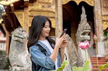 Olumlu, çekici genç Asyalı bir bayan turist Tayland 'da bir tapınağı ziyaret ederken bir nilüfer çiçeğinin fotoğrafını çekmek için akıllı telefonunu kullanıyor. İnsanlar, yaşam tarzı ve kablosuz teknoloji