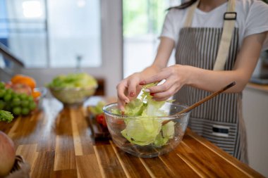 Mutfak masasında salata kasesi hazırlayan, evde sağlıklı yemek yapan önlüklü bir kadının yakın plan görüntüsü. sağlıklı yaşam tarzı, ev hayatı, diyet yemekleri