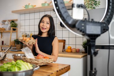 Çekici bir Asyalı kadın yemek blogcusu veya içerik yaratıcısı kamera önünde yemek pişiriyor, konuşuyor ve mutfak stüdyosunda kayıt yaparken özel bir menü için malzemelerini gösteriyor..