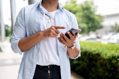 Sıradan kıyafetlerle gezen Asyalı bir adamın kırpılmış görüntüsü şehirde yürürken akıllı telefonunu kullanıyor. Sohbet etmek, cevaplamak, telefonda kaydırmak, e-postaları kontrol etmek, online haberleri okumak.