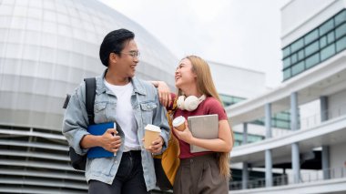 İki neşeli Asyalı genç üniversite öğrencisi, bir erkek ve bir kadın, günlük giysiler içinde, arka planda modern üniversite binalarıyla dışarıda yürürken gülüyor ve eğleniyorlar..