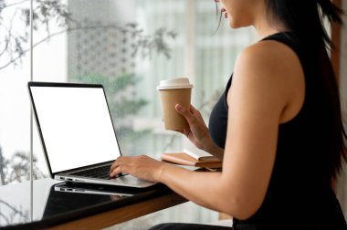 Çekici bir Asyalı kadının minimalist bir kahve dükkanından kahve içerken dizüstü bilgisayar kullanarak uzaktan çalışmasının kırpılmış bir yan görüntüsü. İnsanlar, yaşam tarzı, teknoloji