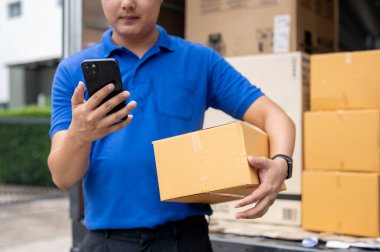 Mavi üniformalı bir Asyalı teslimatçının kesilmiş bir resmi kamyonetinin arkasında duruyor, bir paket taşıyor ve teslimatı yapmadan önce müşterinin adresini akıllı telefonundan kontrol ediyor..