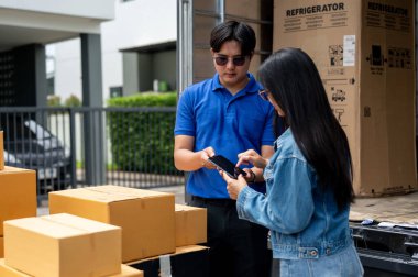 Asyalı bir bayan müşteri paketlerini teslimatçıdan almadan önce akıllı telefonu imzalıyor, evinin önünde duruyor. Nakliye, taşıma, nakliye, ticaret.