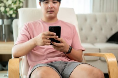 Günlük giysiler içinde Asyalı bir adamın kesilmiş bir görüntüsü oturma odasındaki şezlongda akıllı telefonunu kullanıyor. Sohbet etmek, akıllı telefondan kaymak, cep telefonu uygulaması kullanmak, mesaj göndermek.