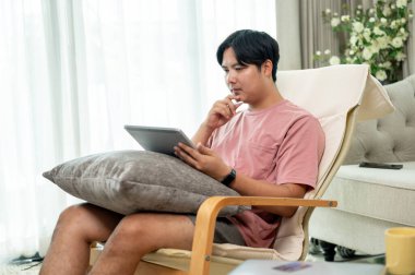 Ciddi bir Asyalı adam günlük giysiler içinde oturma odasındaki şezlongda dijital tablet kullanıyor, evden uzaktan çalışıyor. İnsanlar ve kablosuz teknoloji kavramları