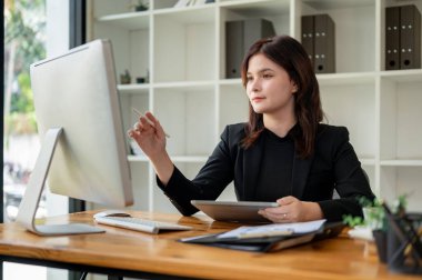 Siyah takım elbiseli güzel, zeki bir iş kadını masasında modern, parlak bir ofiste çalışırken bilgisayar ekranına bir kalemle işaret ediyor..