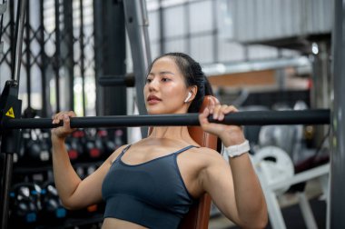 Spor giyimli çekici bir Asyalı kadın spor salonunda ağırlık kaldırıyor ve göğüs ve trisepslerini çalıştırmaya odaklanıyor..