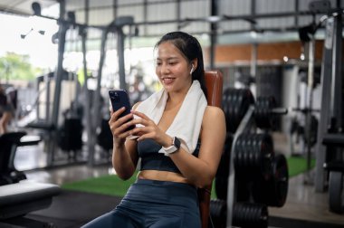 Sportif giyimli neşeli bir Asyalı kadın spor salonundaki antrenmanından sonra ağırlık kaldırma makinesinde dinlenirken akıllı telefonunu kullanıyor. İnsanlar, spor ve sağlıklı yaşam tarzı kavramları