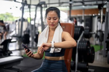Sportif giyimli Asyalı bir kadın spor salonundaki bir bank presinin üzerinde dinlenirken spor sonuçlarına bakarak yanık kalorilerini takip ediyor. gadget ve spor yaşam tarzı kavramları