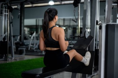 Spor elbiseli Asyalı bir kadının arka plan görüntüsü bir bankta oturuyor, üst sırt kaslarını çalıştırmak için bir kablo çekiyor, spor salonundaki çoklu istasyon makinesinde çalışıyor..