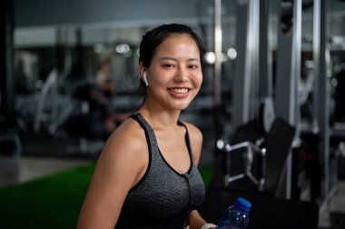 Spor salonunda spor yaparken, spor yaparken kameraya gülümseyen, spor yaptıktan sonra bankta dinlenen güzel, mutlu bir Asyalı kadın. Spor, eğitim, sağlıklı yaşam tarzı, egzersiz