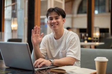 Yakışıklı, pozitif Asyalı genç bir adam bir kahve dükkanından uzaktan çalışıyor, kanepede oturuyor, dizüstü bilgisayarı masanın üstünde, elini sallıyor ve kameraya gülümsüyor. modern yaşam tarzı, teknoloji