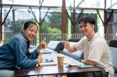 İki pozitif, mutlu Asyalı genç üniversite öğrencisi, bir erkek ve bir kadın, bir kafede ödev yapıyorlar, kameraya gülümsüyorlar ve başparmak işareti yapıyorlar..