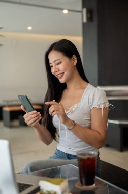 Modern bir kafede otururken akıllı telefonunu kullanan güzel, pozitif Asyalı bir kadın. Metin okumak, sohbet cevaplamak, mesajlar göndermek, telefonda kaydırmak, internette gezinmek
