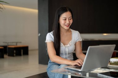 Çekici, gülümseyen Asyalı bir kadın bir kafede çalışıyor, dizüstü bilgisayarı üzerinde çalışıyor. şehir hayatı, yaşam tarzları, kablosuz teknoloji kavramları