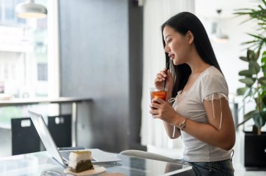Güzel bir Asyalı kadının laptopunda bir şey okuyup, kahve dükkanından uzaktan çalışırken soğuk americano yudumlamasının yan görüntüsü. Modern şehir yaşam tarzı ve kablosuz teknoloji