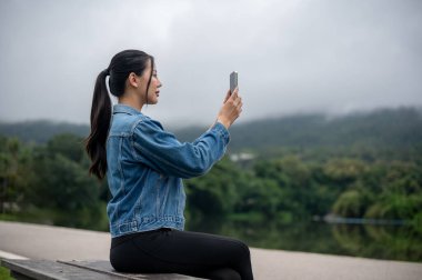 Kot ceketli güzel bir Asyalı kadın çarpıcı bir gölün kenarındaki bankta oturuyor, akıllı telefonunu manzaranın fotoğrafını çekmek için kullanıyor. İnsanlar, yaşam tarzı ve teknoloji