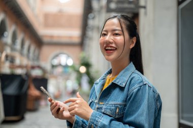 Kot ceketli, güzel, pozitif Asyalı bir kadın arka planda bir binayla dışarıda duruyor, akıllı telefonu tutuyor ve başka tarafa bakarken gülümsüyor. İnsanlar, yaşam tarzı, kablosuz teknoloji