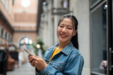 Kot ceketli güzel bir Asyalı kadın arka planda binalar ve mağazalarla dışarıda duruyor, akıllı bir telefon tutuyor ve kameraya gülümsüyor. İnsanlar, yaşam tarzları, kablosuz teknoloji