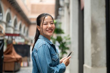 Kot ceketli güzel bir Asyalı kadın arka planda binalar ve mağazalarla dışarıda duruyor, akıllı bir telefon tutuyor ve kameraya gülümsüyor. İnsanlar, yaşam tarzları, kablosuz teknoloji