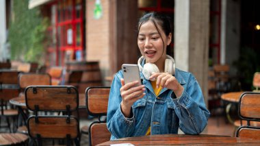 Kot ceketli güzel, pozitif Asyalı bir kadın bir restoranın açık masasında otururken akıllı telefonunu kullanıyor. kısa mesajları okumak, sohbet etmek, mobil bir uygulama kullanmak