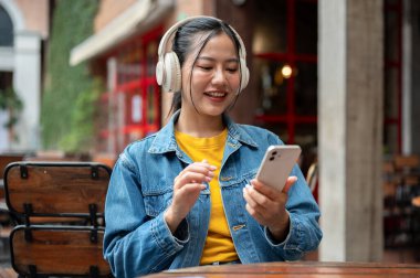 Kot ceketli, güzel, mutlu bir Asyalı kadın bir restoranın açık masasında otururken akıllı telefonunu kullanıyor ve kulaklıklarıyla müzik dinliyor..