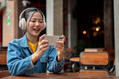 Kot ceketli, kulaklık takan mutlu bir Asyalı kadın, gezici bir oyun oynamaktan ya da bir restoranın açık masasında otururken telefonundan video izlemekten zevk alıyor..