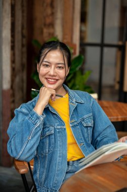 Kot ceketli çekici, pozitif Asyalı bir kadın kahve dükkanında oturmuş kameraya gülümserken kitap okuyor. insanlar ve yaşam tarzı kavramları