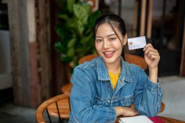 Kot ceketli, çekici, gülümseyen Asyalı bir kadın kahve dükkanında ya da restoranda oturuyor, kredi kartını tutuyor ve kameraya gösteriyor. Nakitsiz ödeme, e-ticaret, bankacılık, finans