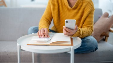 Evdeki kanepede rahat bir kazakla oturan, ev giderlerini ve aylık birikimlerini planlamak ve yönetmek için telefonunu ve hesap makinesini kullanan bir kadının yakın çekimi. insanlar ve finansal kavramlar