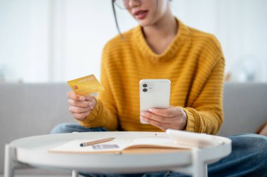Kanepede oturan, elinde akıllı telefonu ve kredi kartıyla internetten alışveriş yapan rahat sarı kazaklı mutlu bir Asyalı kadın. E-ticaret, nakit ödeme, mobil bankacılık.