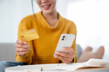 Kanepede oturan, elinde akıllı telefonu ve kredi kartıyla internetten alışveriş yapan rahat sarı kazaklı mutlu bir Asyalı kadın. E-ticaret, nakit ödeme, mobil bankacılık.