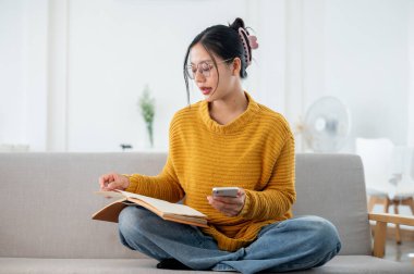 Günlük giysiler içinde güzel bir Asyalı kadın evdeki bir kanepede kitap okumaya odaklanıyor. ev hayatı, boş zaman, yaşam tarzları