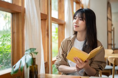 Sıradan giysiler içinde çekici bir Asyalı kadın minimalist bir kahve dükkanının penceresinin yanındaki masaya oturur, pencereden dışarı bakar ve kafede kitap okumaktan keyif alırken hayal kurar..