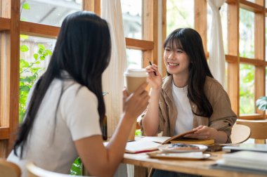 İki neşeli genç Asyalı üniversite öğrencisi minimalist bir kafede konuşmaktan ve okumaktan keyif alıyorlar. İnsanlar, yaşam tarzları, üniversite hayatı, boş zaman