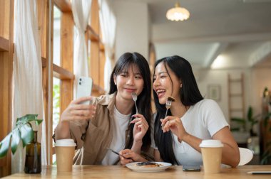 İki güzel ve mutlu Asyalı kadın bir kafede takılırken akıllı telefonlarıyla fotoğraf çekmekten zevk alıyorlar. İnsanlar, yaşam tarzları, kablosuz teknoloji