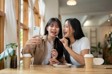 İki güzel ve mutlu Asyalı kadın bir kafede takılırken akıllı telefonlarıyla fotoğraf çekmekten zevk alıyorlar. İnsanlar, yaşam tarzları, kablosuz teknoloji