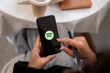 Chiang Mai, Tayland - Ekim 03 2024: Ekranda Spotify logosu olan iPhone tutan bir kadının yakın çekimi. Kafede otururken akıllı telefonunda Spotify uygulamasını kullanan bir kadın..