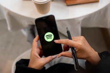 Chiang Mai, Tayland - Ekim 03 2024: Ekranda Spotify logosu olan iPhone tutan bir kadının yakın çekimi. Kafede otururken akıllı telefonunda Spotify uygulamasını kullanan bir kadın..
