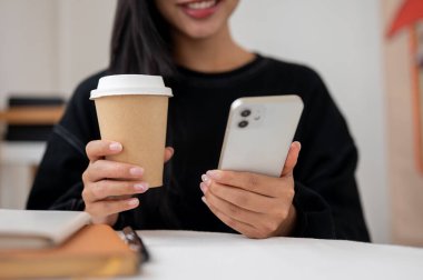 Siyah kazaklı, çekici, pozitif Asyalı bir kadının kapalı bir masada ya da kafede akıllı telefonunda mesaj okurken eğlendiği yakın plan bir fotoğraf. İnsanlar, yaşam tarzları, kablosuz teknoloji