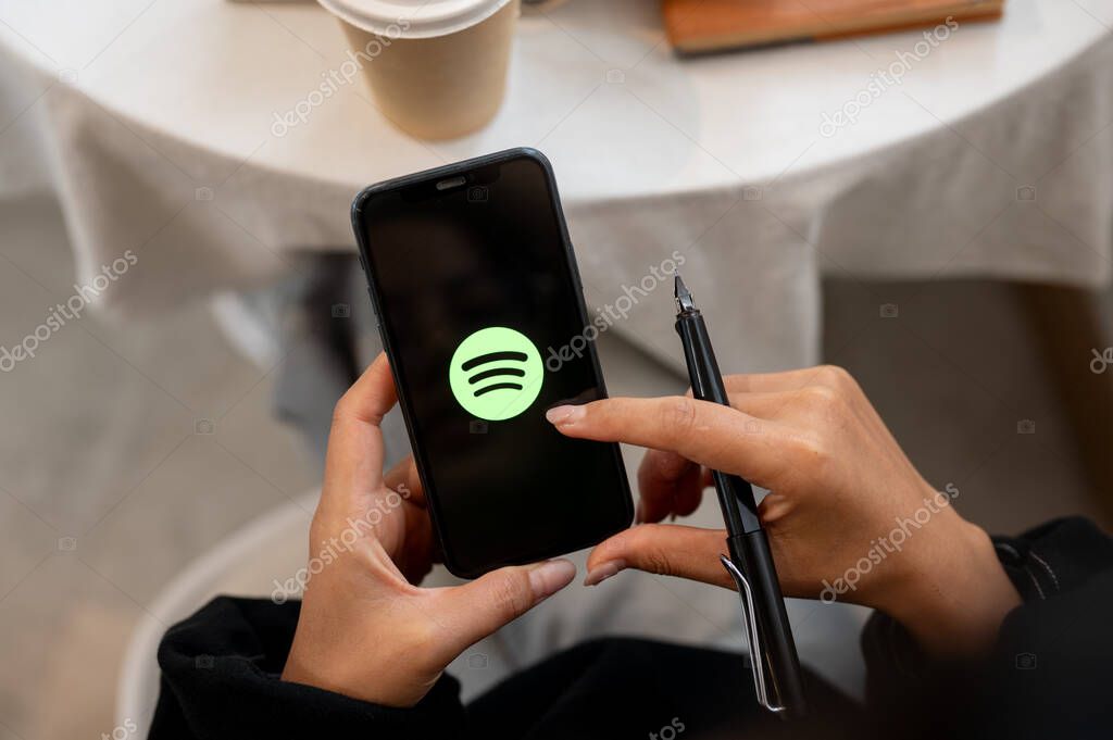 Chiang Mai, Thailand - Oct 03 2024: A close-up of a woman holding an iPhone with the Spotify logo on the screen. A woman using the Spotify application on her smartphone while sitting in a cafe.